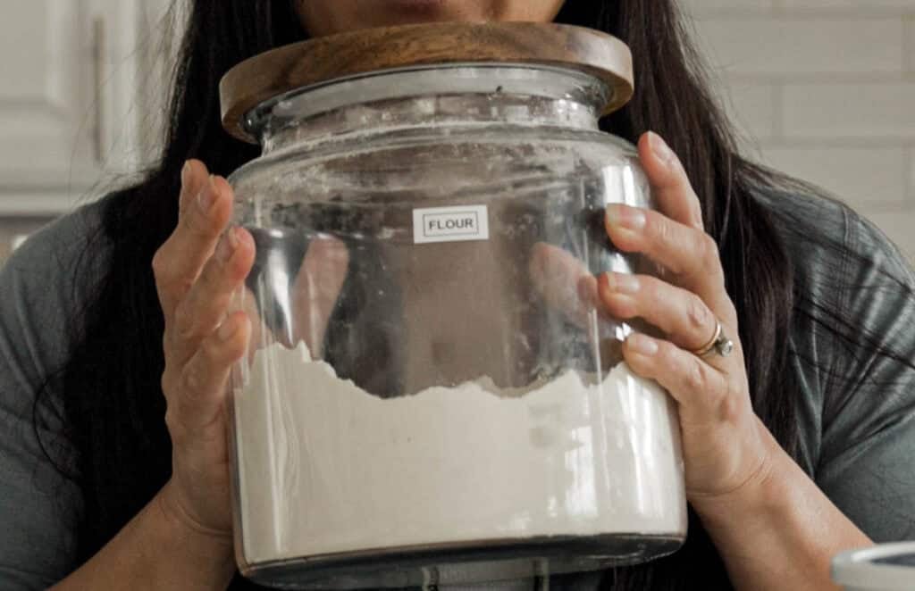 A person holding a large glass jar labeled Flour with a wooden lid, partially filled with flour. The person is wearing grey long sleeves and has long dark hair, standing in a kitchen with a tiled backsplash in the background.